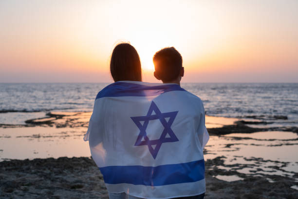 Teenagers, young woman and man with the Flag of Israel draped over their shoulders at the sunset over the sea in israel. Friendship in childhood silhouette
